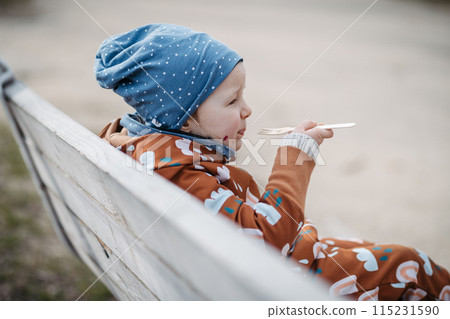Cute toddler girl eating lunch in park, sitting on bench. Picnic in park during cold spring day. 115231590
