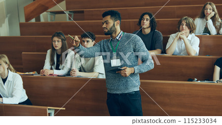 Young teacher, tutor leading lesson in university auditorium, paying attention of students, pointing on blackboard. Multiracial students in classroom. Young teacher, tutor leading lesson in university auditorium, paying attention of students, pointing on blackboard. Multiracial students in classroom. 115233034