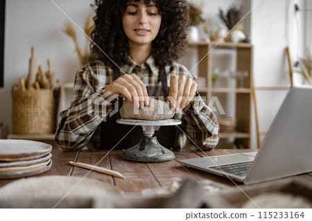 Woman creating pottery in her home studio with laptop Woman creating pottery in her home studio with laptop 115233164