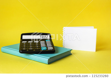 calculator with the word TRUST on the display of a calculator with a notepad on a yellow background 115233260