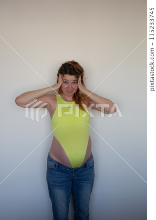 A joyful pregnant woman in a yellow bodysuit and jeans, smiling with excitement and anticipation 115233745