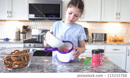 With focused attention, this budding culinary artist dips pretzels into a pot of melted chocolate, creating sweet delights in the warmth of a well-lit home kitchen. 115233788