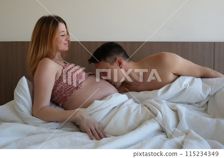 A loving couple in bed, he kisses her pregnant belly, sharing a tender moment 115234349