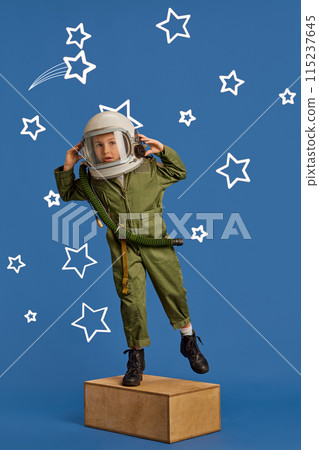 Portrait of young dreamer, little boy in pilot's uniform and helmet aiming high among stars against blue background. 115237645