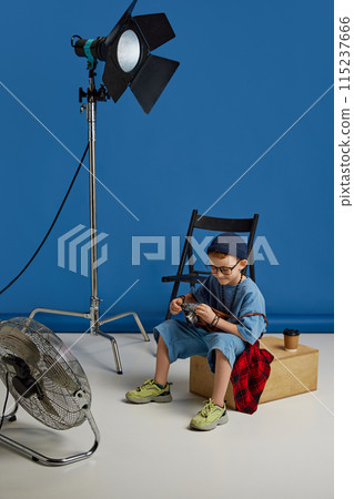 Boy with camera and coffee cup, sitting near photo set equipment, dreaming of career in photo production against blue background. Boy with camera and coffee cup, sitting near photo set equipment, dreaming of career in photo production against blue background. 115237666