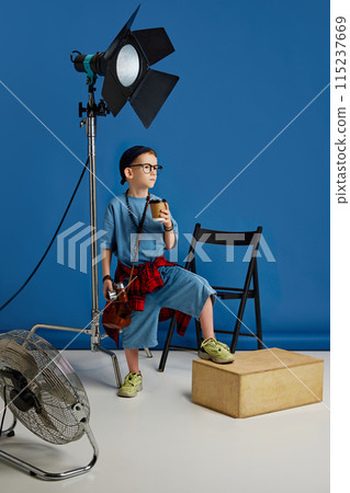 Portrait of confident young boy, dressed casual attire, stands in studio near photographer equipment and drinks coffee against blue background. Portrait of confident young boy, dressed casual attire, stands in studio near photographer equipment and drinks coffee against blue background. 115237669