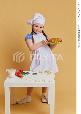 Little girl with chef's hat, serving delicious fresh baked croissant, dreaming of culinary future against beige studio background. Little girl with chef's hat, serving delicious fresh baked croissant, dreaming of culinary future against beige studio background. 115237686