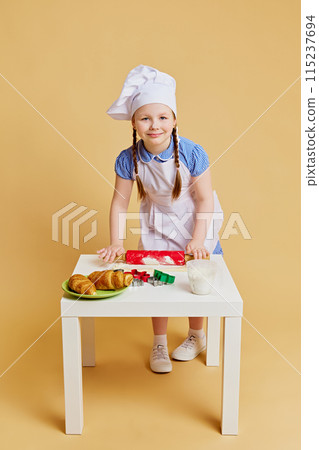 Portrait of little girl dressed as chef, using cookie rolling pin on dough, dreams of future in baking against beige studio background. 115237694