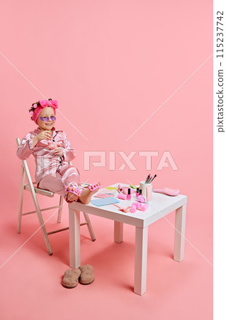 Girl enjoying spa day, in pajamas, with beauty products and eye patches under eyes against pastel pink studio background. 115237742