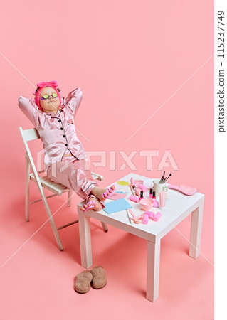 Little girl in pajamas enjoys spa day with cucumber slices on their eyes and beauty products on table against pastel pink studio background. 115237749