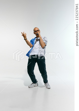 Young man dressed in split-patterned blue and white shirt with glasses, sings energetically into microphone against white studio background. 115237841