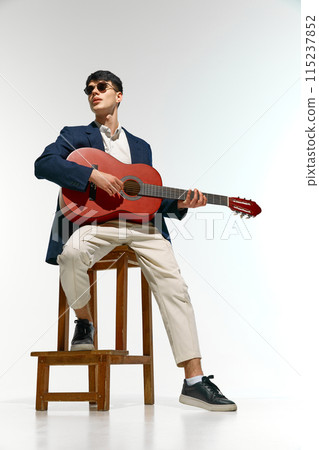 Fashionable man dressed in blue blazer and light trousers, complete with sunglasses, plays strumming guitar while seated against white studio background. 115237852