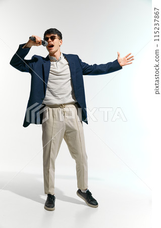 artistic man, vocalist dressed casual attire with sunglasses performing, singing with microphone against white studio background. 115237867