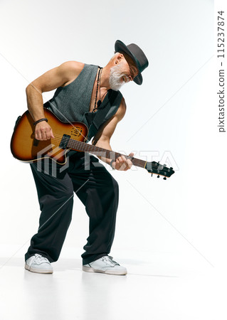 Man with beard, wearing gray sleeveless shirt and stylish hat, deeply engrossed in playing electric guitar against white studio background. Man with beard, wearing gray sleeveless shirt and stylish hat, deeply engrossed in playing electric guitar against white studio background. 115237874