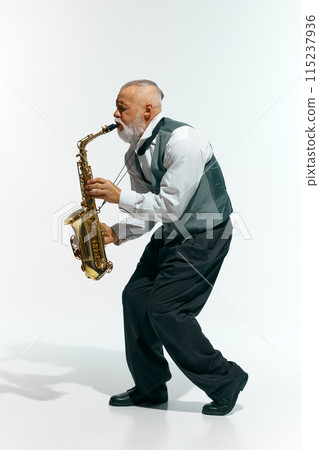 Jazzy vibes. Bearded musician in sleeveless gray vest and black pants plays saxophone energetically against white studio background. Jazzy vibes. Bearded musician in sleeveless gray vest and black pants plays saxophone energetically against white studio background. 115237936