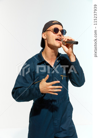 Portrait of young man, musician wearing overalls with sunglasses singing with microphone against white studio background. Portrait of young man, musician wearing overalls with sunglasses singing with microphone against white studio background. 115237939