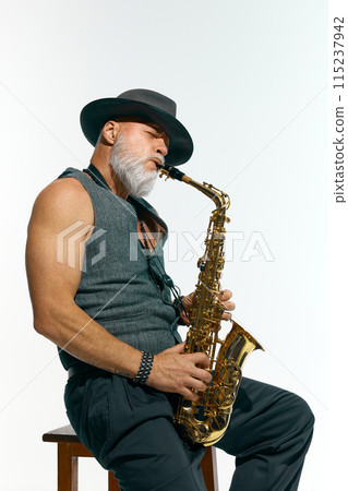 Bearded man in sleeveless gray vest and black hat playing jazz, golden saxophone against white studio background. Bearded man in sleeveless gray vest and black hat playing jazz, golden saxophone against white studio background. 115237942