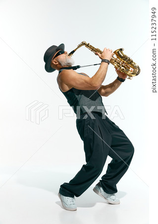 Bearded man wearing sleeveless gray vest, black pants, and stylish hat passionately plays golden saxophone against white studio background. 115237949