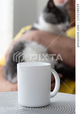Woman sits at a kitchen table with a cat in her hands and drinks tea with cats hair on a white cup. Moulting cat. Selective focus. 115238034