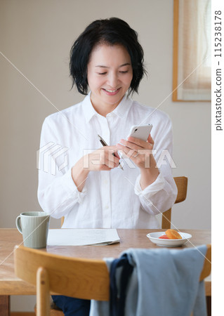 A middle-aged woman searching for information on her smartphone and taking notes in the living room 115238178