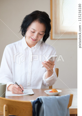 A middle-aged woman searching for information on her smartphone and taking notes in the living room 115238180