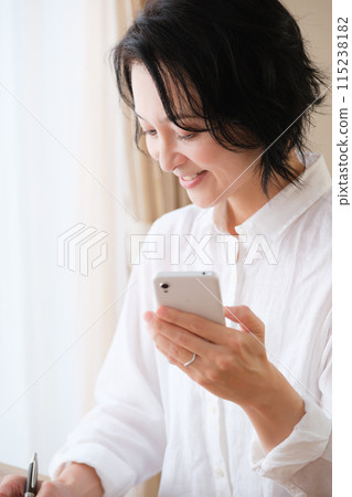 中年婦女在客廳用智慧型手機搜尋資訊並做筆記 中年婦女在客廳用智慧型手機搜尋資訊並做筆記 115238182