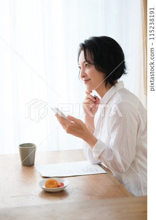 A middle-aged woman searching for information on her smartphone and taking notes in the living room 115238191
