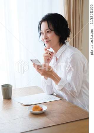A middle-aged woman searching for information on her smartphone and taking notes in the living room 115238196