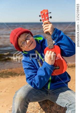A boy is seated on a rock, playing a ukulele under the sunny sky 115239403