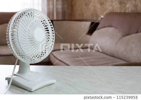 Tabletop compact white plastic fan with included blades disperses cold air throughout the home room against the backdrop of the sofa on hot day Tabletop compact white plastic fan with included blades disperses cold air throughout the home room against the backdrop of the sofa on hot day 115239958