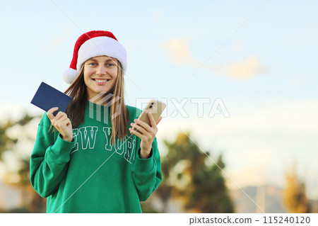 Season winter vacations. Smiling attractive woman in Santa Claus hat holds passport and smartphone for online booking ticket, hot tours for travels on Christmas and New Year holiday. Copy space Season winter vacations. Smiling attractive woman in Santa Claus hat holds passport and smartphone for online booking ticket, hot tours for travels on Christmas and New Year holiday. Copy space 115240120