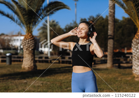 Young female athlete 20-30 years takes a break from her workout in sportswear and earphones outdoors on palm trees backdrop. Woman is happily smiling, combined physical fitness with joy and wellbeing. 115240122