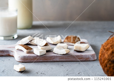 Fresh coconut pieces on a wooden board with a glass and jar of coconut milk in the background, showcasing natural ingredients. 115241001