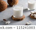 Two glasses of fresh coconut milk on wooden coasters, accompanied by sliced coconut pieces and whole coconuts on a textured surface. 115241003