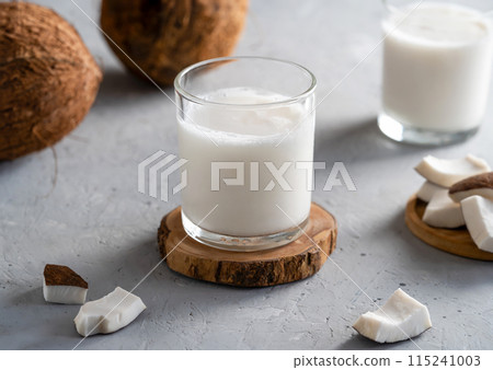 Two glasses of fresh coconut milk on wooden coasters, accompanied by sliced coconut pieces and whole coconuts on a textured surface. Two glasses of fresh coconut milk on wooden coasters, accompanied by sliced coconut pieces and whole coconuts on a textured surface. 115241003