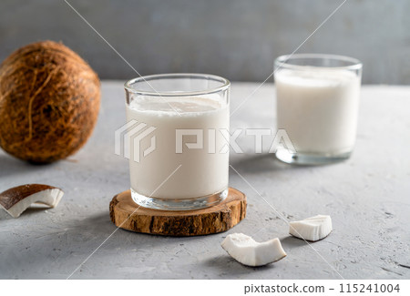 Two glasses of fresh coconut milk on wooden coasters with sliced coconut pieces and a whole coconut on a textured gray surface. Two glasses of fresh coconut milk on wooden coasters with sliced coconut pieces and a whole coconut on a textured gray surface. 115241004