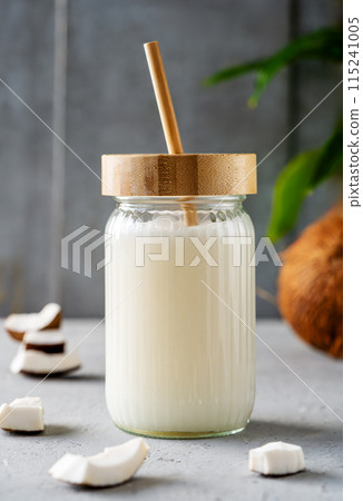 A jar of fresh coconut milk with a bamboo straw, surrounded by coconut pieces and a whole coconut on a textured surface. A jar of fresh coconut milk with a bamboo straw, surrounded by coconut pieces and a whole coconut on a textured surface. 115241005
