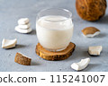 A glass of fresh coconut milk placed on a wooden coaster, surrounded by coconut pieces on a textured gray surface. 115241007