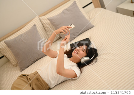 Smiling young woman listening to music with headphones and using smartphone while lying in bed 115241765
