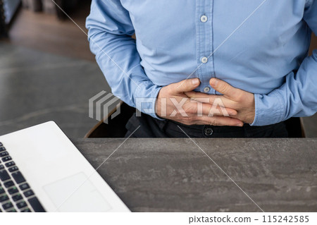 Businessman in blue shirt experiencing stomach pain while working on laptop at office desk, highlighting health issues related to stress or digestion. 115242585