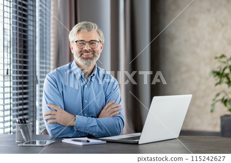 Confident senior businessman wearing glasses and a blue shirt working at his desk with a laptop in a modern office setting, smiling at the camera. 115242627