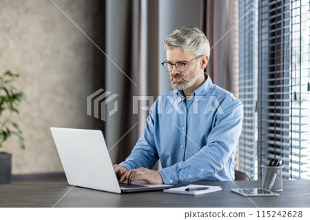 Mature professional man in blue shirt working on a laptop in a modern office setting with natural light and focus on his work. 115242628