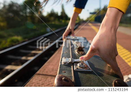 A girl fastens the zipper on an old travel suitcase on a train platform one summer afternoon during a planned train trip. A girl fastens the zipper on an old travel suitcase on a train platform one summer afternoon during a planned train trip. 115243194