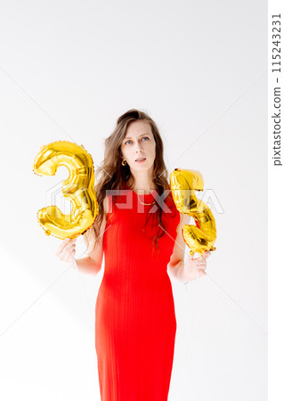 Beautiful woman with balloons and the numbers 35 is photographed in the studio in a red dress. Beautiful and feminine woman celebrates her 35th birthday 115243231