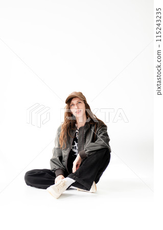 Young woman in a bomber jacket, jeans and a cap with braces poses while sitting in the studio, on a white background Young woman in a bomber jacket, jeans and a cap with braces poses while sitting in the studio, on a white background 115243235