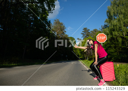 A young adult stands on the side of the road with a pink suitcase and tries to stop the other cars to get a free ride to the holiday destination. 115243329