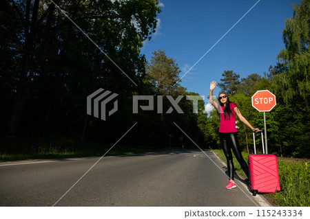 A young adult stands on the side of the road with a pink suitcase and tries to stop the other cars to get a free ride to the holiday destination. A young adult stands on the side of the road with a pink suitcase and tries to stop the other cars to get a free ride to the holiday destination. 115243334