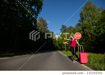 A young adult stands on the side of the road with a pink suitcase and tries to stop the other cars to get a free ride to the holiday destination. A young adult stands on the side of the road with a pink suitcase and tries to stop the other cars to get a free ride to the holiday destination. 115243342