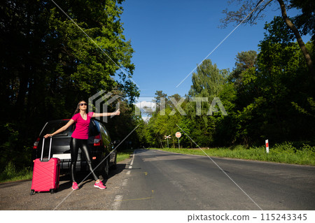 A young girl stands on the road in a green forest next to a broken car and asks for help, stopping other cars. 115243345