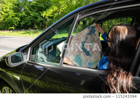 A young girl sits in a car parked on the side of the road in the woods and reads a map to find the right route to its destination. A young girl sits in a car parked on the side of the road in the woods and reads a map to find the right route to its destination. 115243394